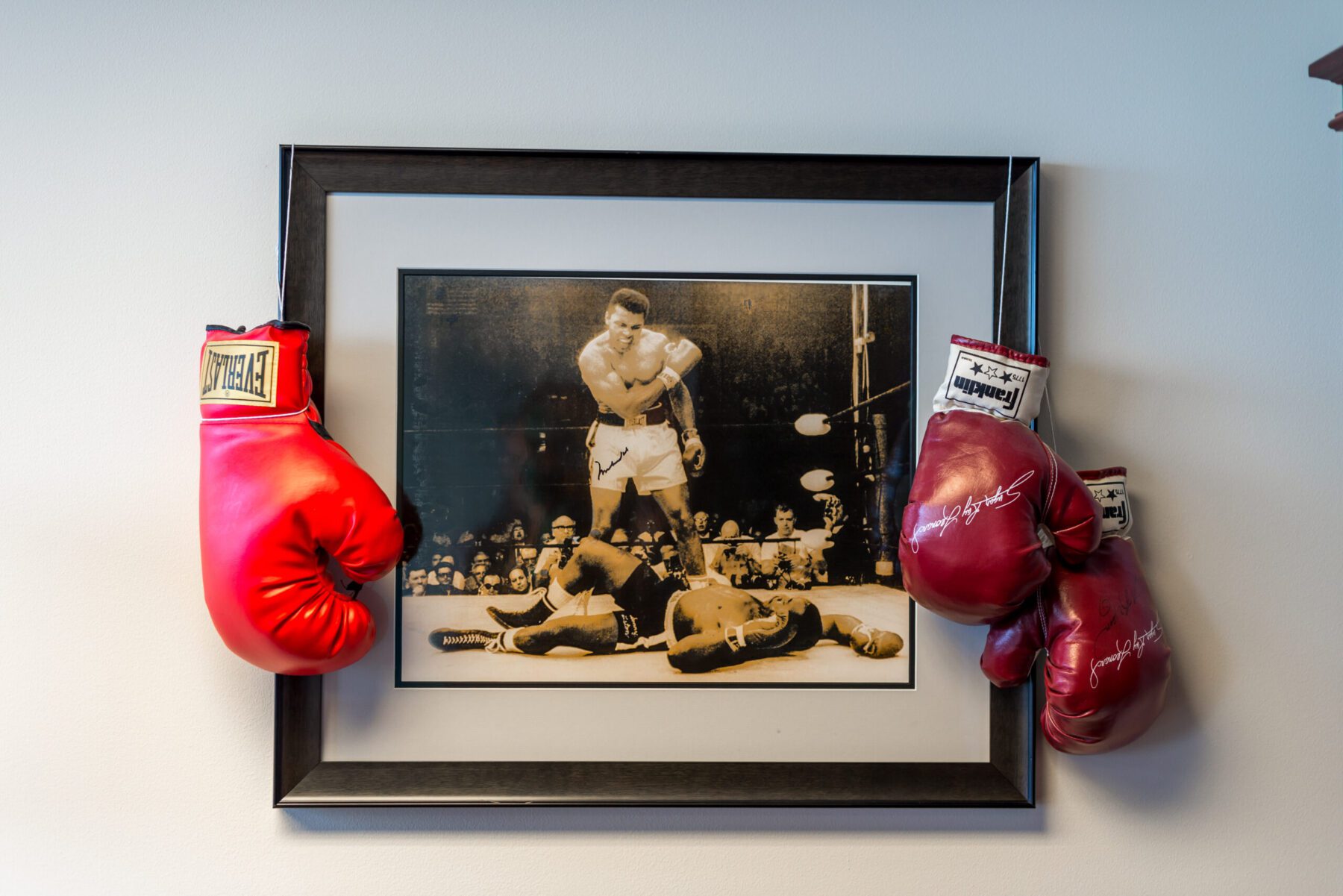 A sepia toned photo of Muhammad Ali just after knocking out his opponent. Two pairs of boxing gloves hang from the black frame.