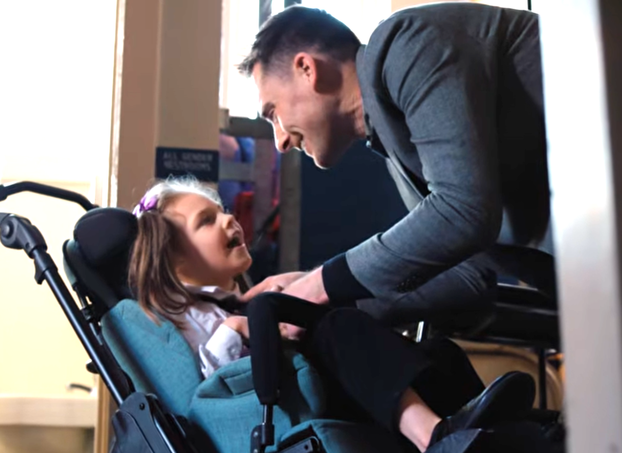 Still from the AT&T video: a little girl in a reclined wheelchair and her father leaning in, both smiling at each other.
